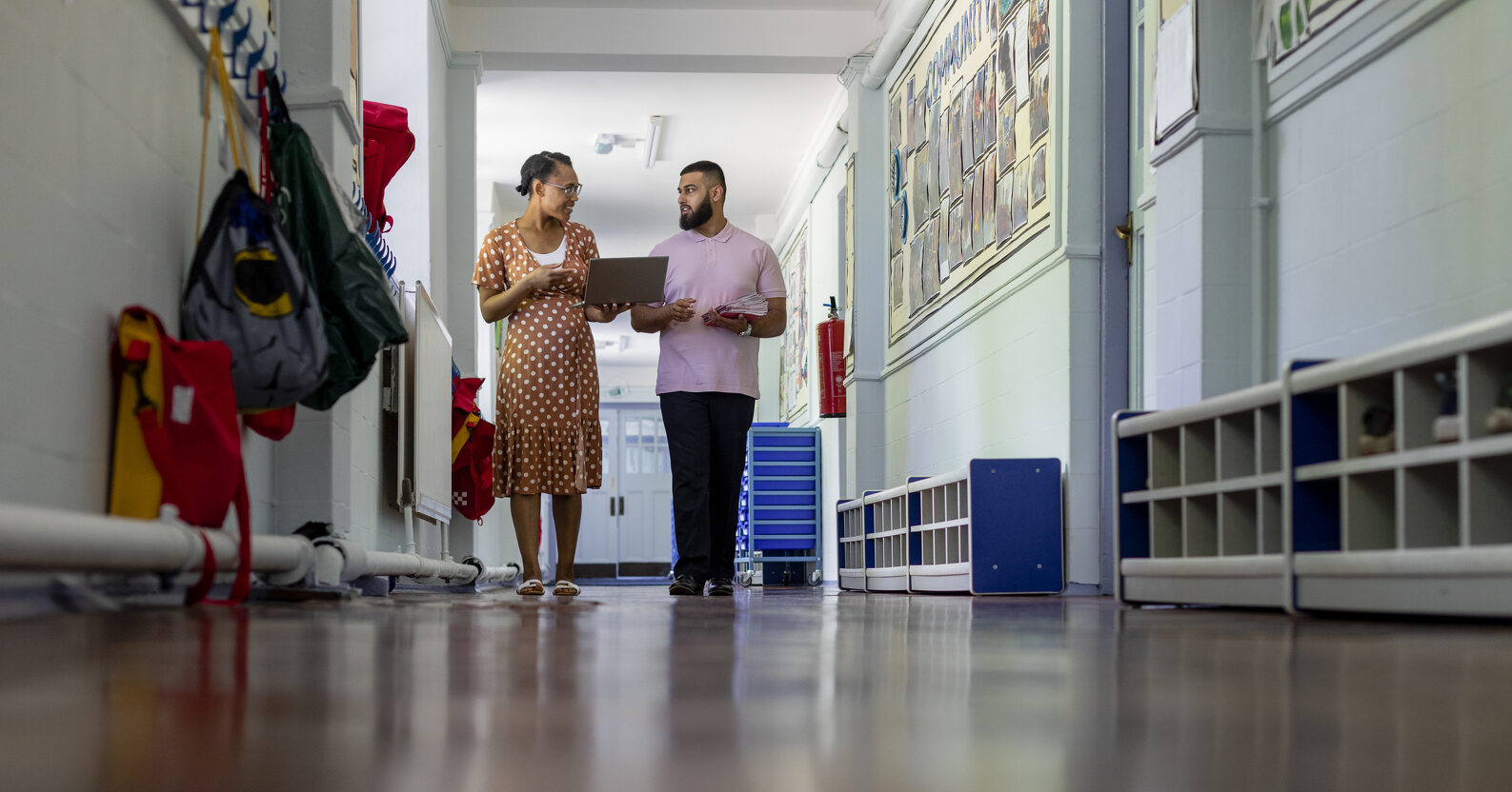 Primary school teachers looking over lesson materials together in the North East of England while walking down a hallway. 2023/02/iStock-1346505098-e1691098798638.jpg 